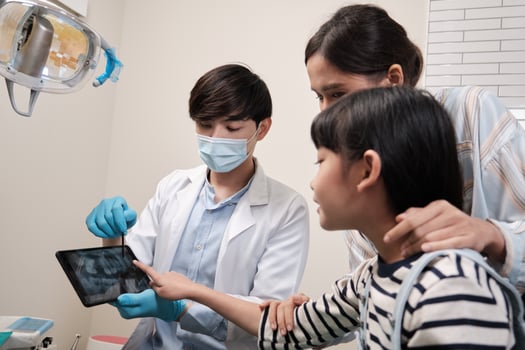 A professional Asian male pediatric dentist explains oral and teeth X-ray result to girl and mom in dental clinic, mouth dentistry hygiene examination, and orthodontic healthcare work in kid hospital. photo
