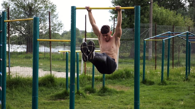 Athletic man trains abdominal muscles raising legs on horizontal bar at sports ground. Young sportsman exercising on pull-up bar outdoor. Sporty athlete working out at nature. Healthy lifestyle photo