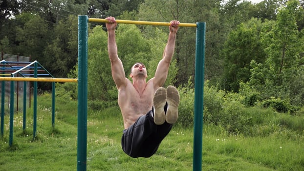 Athletic man trains abdominal muscles raising legs on horizontal bar at sports ground. Young sportsman exercising on pull-up bar outdoor. Sporty athlete working out at nature. Healthy lifestyle. photo
