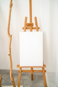 Wooden easel with blank canvas on white wall background. Wooden decor element nearby. Vertical photo