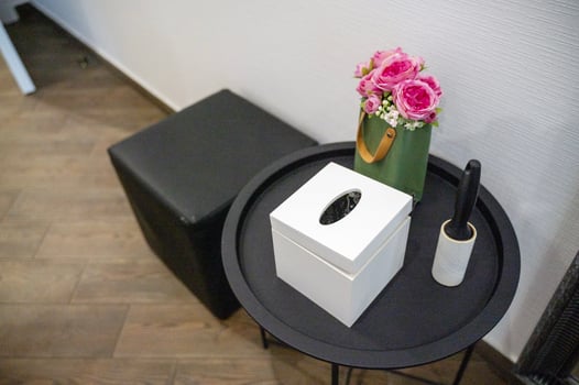 round black metal table next to it pouf. on the table are a box for napkins, decorative flowers, a brush for cleaning clothes. top view. on the floor are wood-like tiles. the walls are white. photo