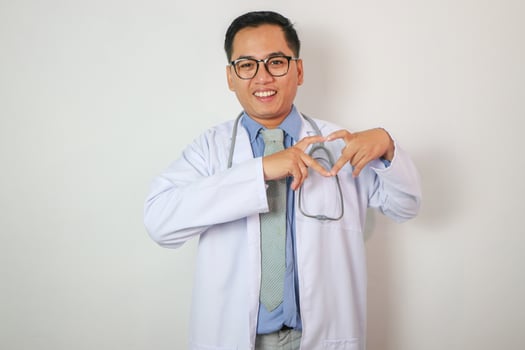 Portrait of Middle aged, handsome, Asian, doctor, on duty at hospital stethoscope around his neck, proudly making heart shape sign for love and support, on isolated white background photo