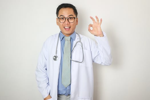 Happy middle aged asian male therapist in white coat, protective gloves with stethoscope showing ok sign, isolated on gray background. Doctor recommendation and advice for health care and medicine photo