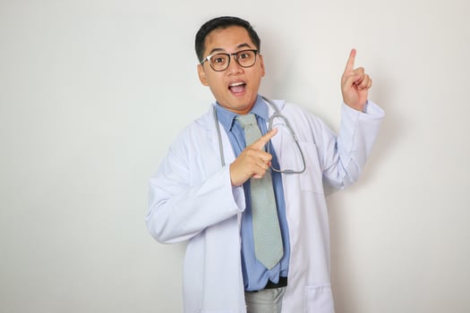 Smiling professional young Asian male doctor or nurse wearing a blue uniform pointing finger up isolated on white background. Healthcare medicine concept photo