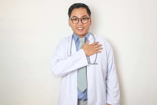 A young Asian man wearing a white coat puts his hand on his chest in front of a white background. Medical doctor. Scientist. photo