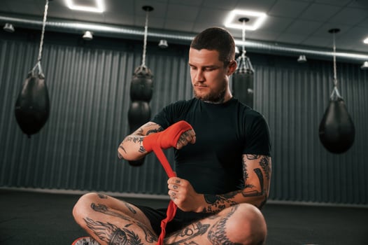 Sitting and wearing red colored hand wraps. Young tattooed man is practicing boxing indoors in the gym. photo