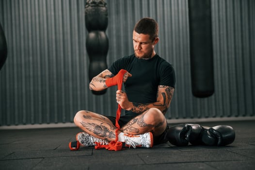 Sitting and wearing red colored hand wraps. ,Young tattooed man is practicing boxing indoors in the gym photo