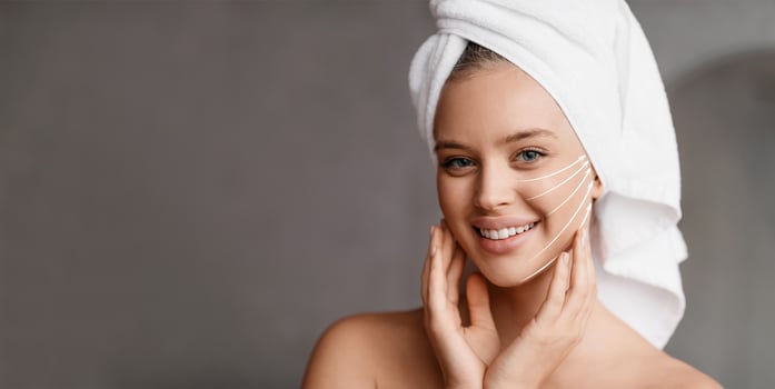 A woman with a towel on her head smiles while pampering her face. She gently massages her skin, embracing self-care and anti-aging practices in a soothing environment, copy space photo