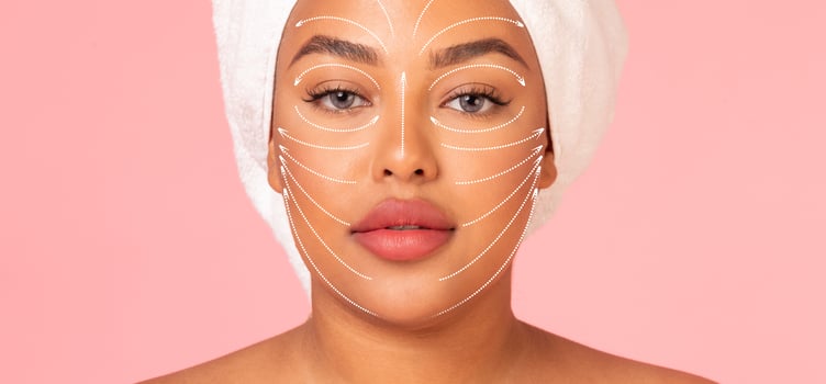 African American woman relaxes with a facial massage focused on rejuvenation and anti-aging techniques. She is wrapped in a towel, highlighting her skin care routine against a soft pink background. photo