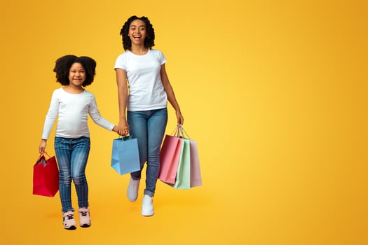 Overjoyed black woman and her cute little daughter holding shopping bags and walking together over pink background in studio, copy space photo