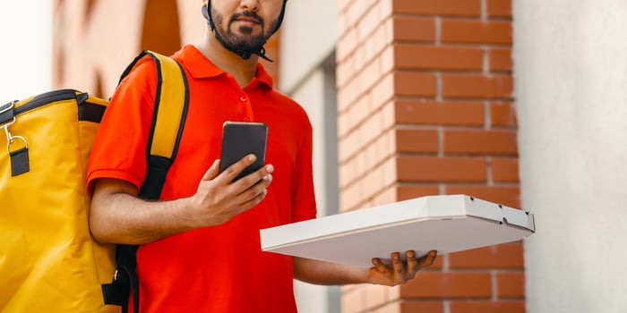 Fast delivery of pizza by courier in city. Serious man in helmet and yellow bag, holds cardboard box with food and watches at smartphone, near front door, copy space photo