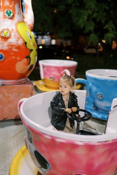 Little girl sits holding the handlebars of a cup-shaped carousel and looks away photo