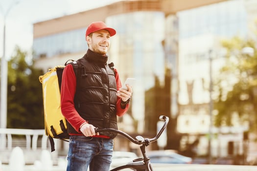 Food Delivery. Courier Guy Holding Smartphone Delivering Lunches Standing With Bike Near Business Center In City. Copy Space photo
