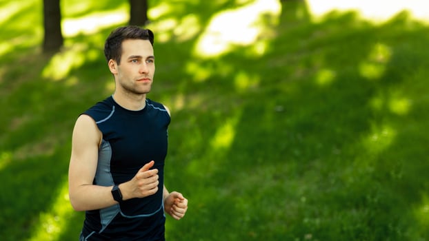 Summer sport. Attractive guy in sportswear with fitness tracker runs in park with green grass, free space, outdoor photo