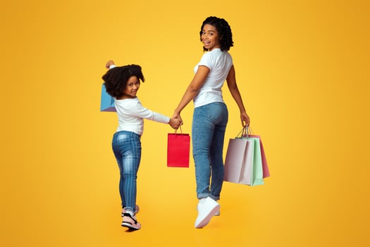 Seasonal Sales. Young african mother and her cute little daughter walking with shopping bags in hands and turning at camera, pink studio background photo