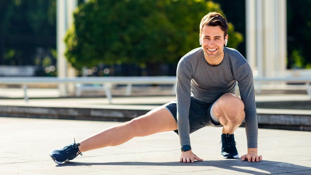 Warm up before run in park. Young smiling man in sports uniform doing leg exercises, outdoor, free space photo