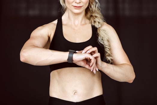 Fitness trainer and workout result. Adult woman in sportswear with fitness tracker and slim belly checks pulse on black background, copy space photo