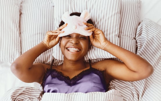 Healthy lifestyle, rest and beauty. Lazy cheerful millennial african american female takes off sleep mask and looks at camera, lies on bed in bedroom, top view, close up, panorama, empty space photo