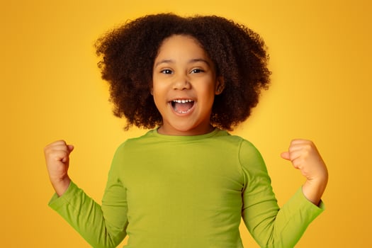 Joyful little black girl celebrating success with raised fists, posing at camera over yellow studio background, free space photo