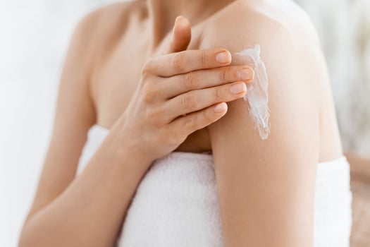 Closeup of young woman wrapped in towel applying body lotion on shoulder after shower, copy space photo