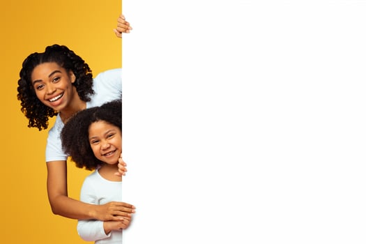 Family Offer. Happy African American Mother And Preschool Daughter Peeking Out Of Blank White Board With Free Space For Your Ad Over Pink Background photo