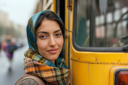 A young Middle-Eastern woman with a warm smile, wearing a colorful scarf, stands beside a yellow bus on a rainy day. photo