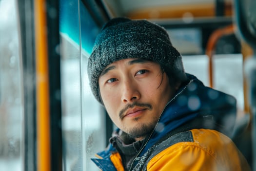A young Asian man gazes thoughtfully out of a bus window during a snowy day, wearing a cozy beanie and a warm jacket. photo