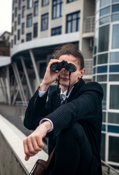 Portrait of a young man against the backdrop of modern architecture, he is looking through binoculars. He has long hair, he is wearing a suit. Front view, close-up. Relaxed, peaceful, thoughtful, happy photo