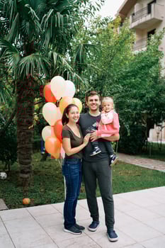 Smiling mom hugging dad with a little girl in his arms while standing in the garden near balloons photo