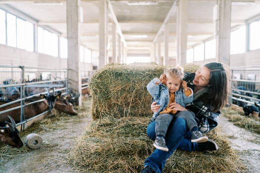 Smiling mom with little girl on her lap sitting on haystack near goat pens at farm photo