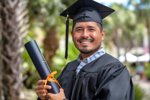 Happy Hispanic male graduate in cap and gown, proudly holding diploma with orange ribbon against a tropical backdrop. photo