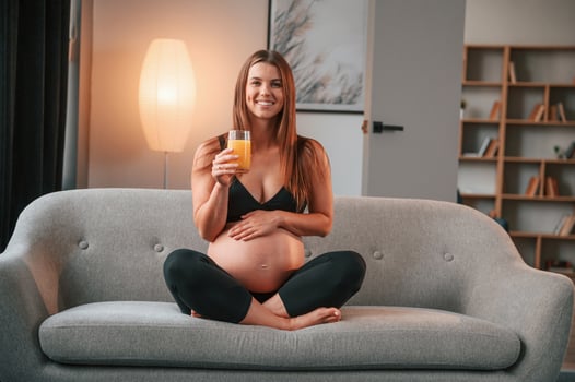 Sitting on the sofa and drinking orange juice. Beautiful pregnant woman is indoors at home photo