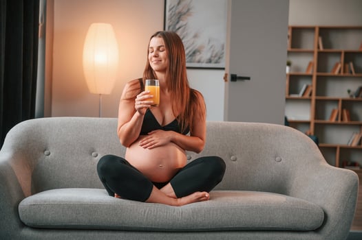 With orange juice in hand.. On the sofa. Beautiful pregnant woman is indoors at home photo