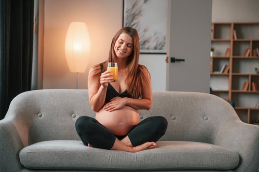 With orange juice in hand. .On the sofa. Beautiful pregnant woman is indoors at home photo