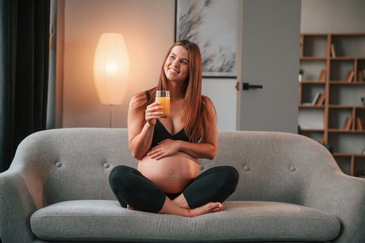 With orange juice in hand. On the sofa. Beautiful pregnant woman is indoors at home photo