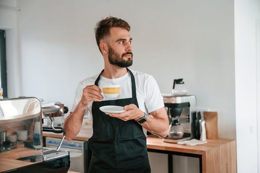Standing and holding cup. Cafe worker ,in white shirt and black apron is indoors photo