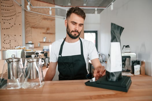 Measuring weight of the package with raw coffee. Cafe worker in white shirt and black apron is indoors photo