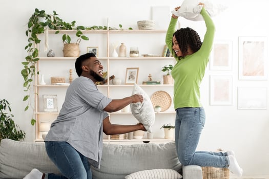 A joyful black couple engages in pillow fight, laughing and playfully challenging each other. The cozy living room is filled with plants and decorative items, enhancing the cheerful atmosphere. photo
