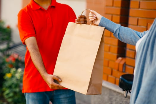 Online shopping, delivery food from restaurants on bike, meal order. Millennial male courier in orange t-shirt gives to woman client eco paper bag near house panorama, empty space, cropped, outdoor photo