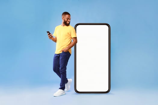 A millennial African American man stands casually with a smartphone in hand, smiling at the screen. He is dressed in a bright yellow shirt and blue pants, with a large blank phone beside him. photo