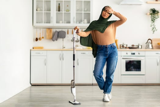 A young woman wearing a headscarf stands in her clean kitchen, looking slightly tired as she takes a break from house cleaning. She uses a steam mop on the floor, showing dedication to her home. photo