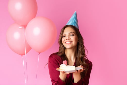 A joyful woman wearing a party hat holds a plate of cake, smiling against a bright pink background with balloons. The atmosphere radiates warmth and celebration during a birthday party. photo