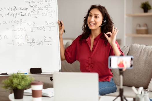 A young middle eastern woman is tutoring students remotely from her cozy living space. She stands next to a whiteboard filled with math equations, smiling and using a headset. photo