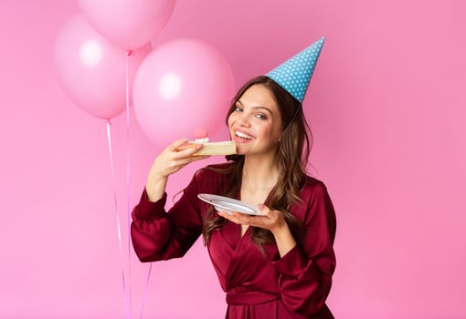 A woman dressed in a stylish outfit smiles while holding a plate with a slice of cake, celebrating with pink balloons in the background. The cheerful mood is highlighted by her festive hat. photo
