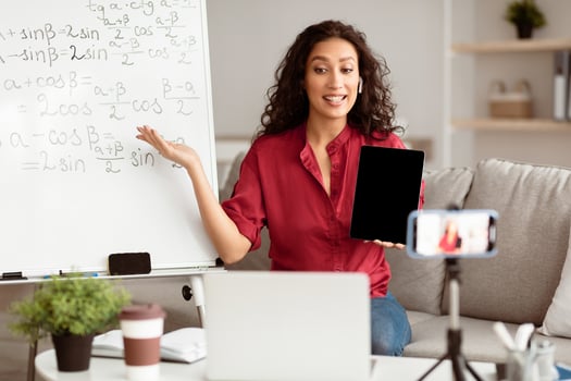 A young Middle Eastern teacher engages students through an online platform. She uses a tablet while explaining concepts in front of a whiteboard filled with equations. photo