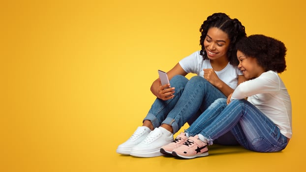 Smiling african girl and her mom sitting on floor with smartphone, watching or cartoons from internet, free space photo