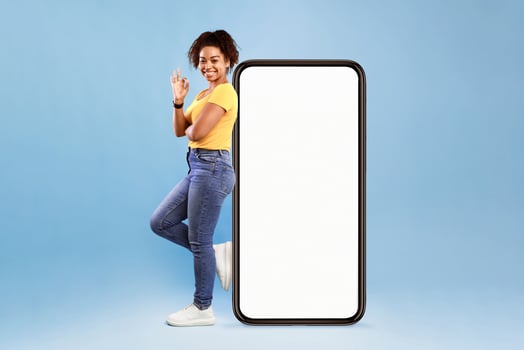 A cheerful African American woman stands confidently beside a large smartphone, wearing a yellow top and jeans. The bright blue backdrop enhances her vibrant and lively personality. photo