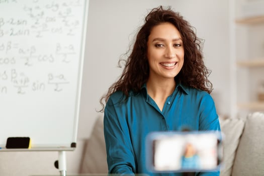 A young Middle Eastern woman conducts an online teaching session, smiling at the camera. She is dressed in a blue shirt and surrounded by a comfortable environment with educational materials. photo
