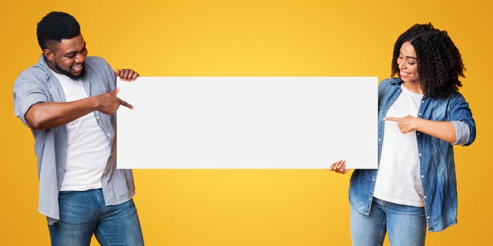A happy couple stands side by side, pointing at a large blank sign. They are smiling and wearing casual clothing, showcasing their enthusiasm against a vibrant yellow backdrop. photo