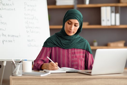 A Middle Eastern woman wearing hijab is engaged in online tutoring from her home office. She takes notes while using her laptop, dressed in comfortable attire. photo